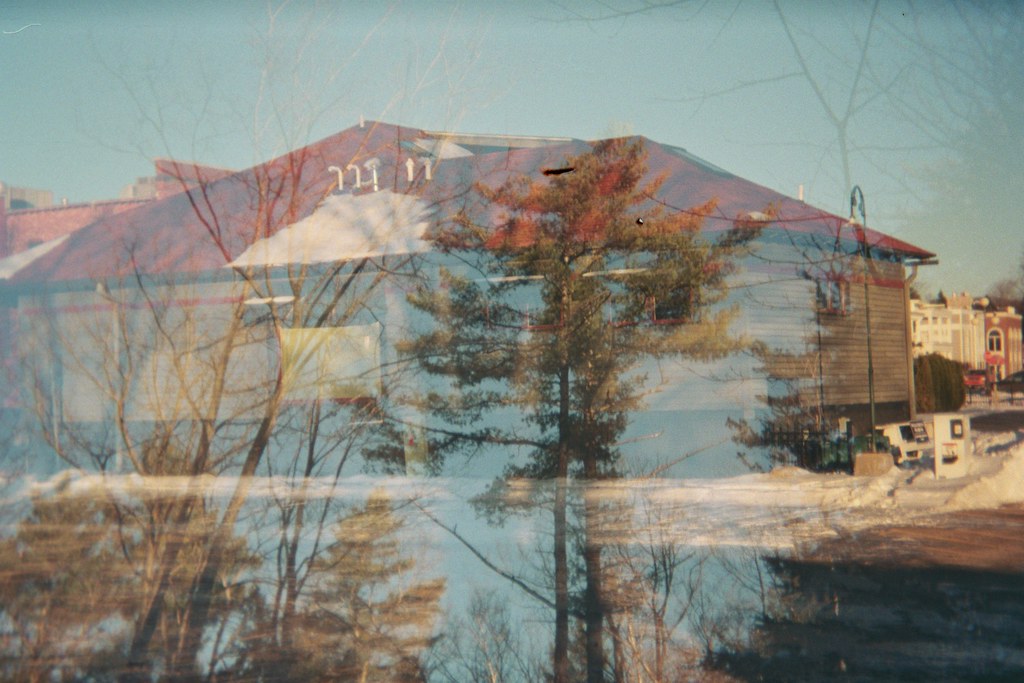 Double-exposure 35mm film photograph layering bare pine trees over a snow-covered building with a red roof.