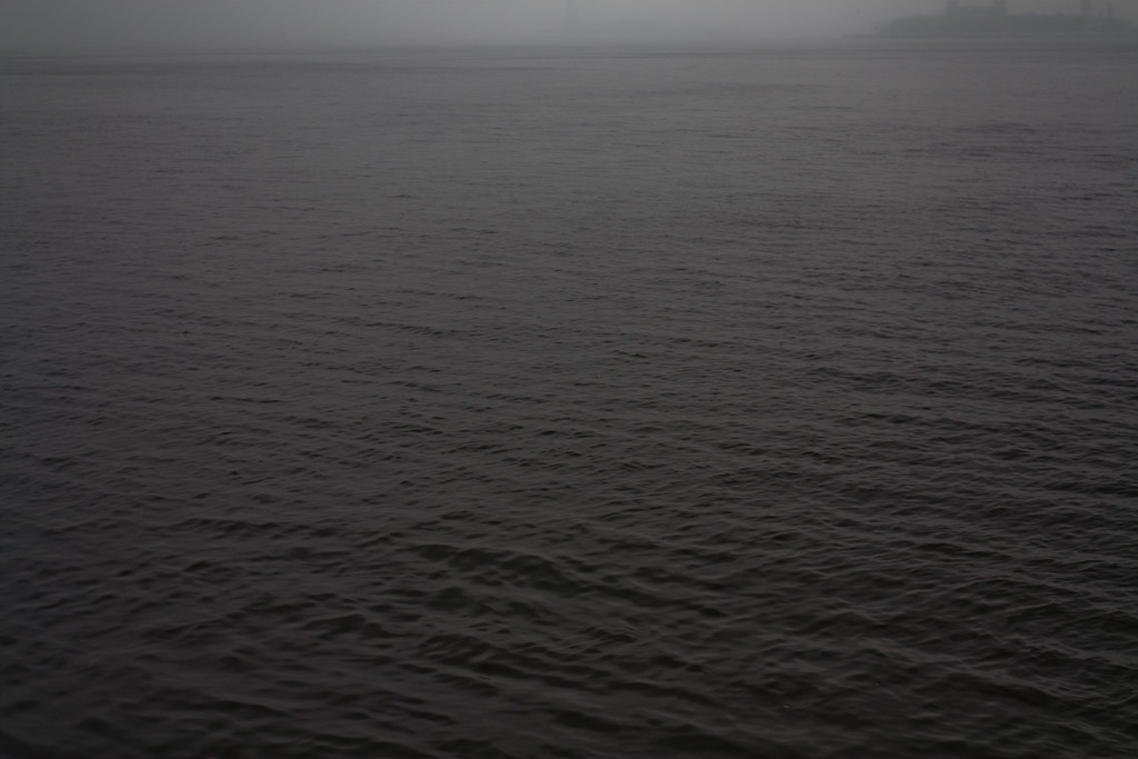 Choppy gray coastal water under dense fog and heavy mist with faint silhouettes of land along the distant horizon.