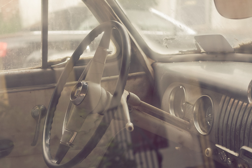 Dusty car window showing a vintage two-spoke steering wheel, column shifter, and chrome dashboard dials.