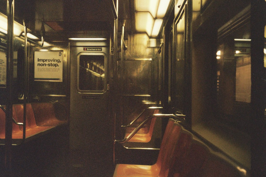 Empty NYC subway car with orange bucket seats, steel poles, and an 'Improving, non-stop.' transit ad.