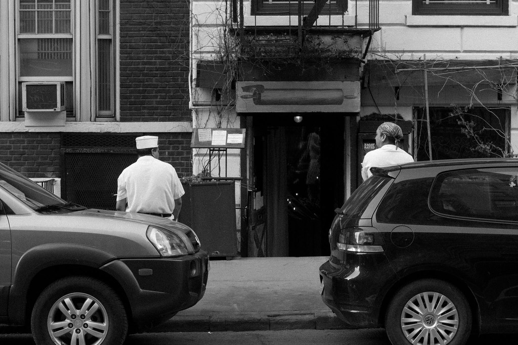 Two restaurant cooks in white taking a break outside an entrance marked by a rolling pin sign.