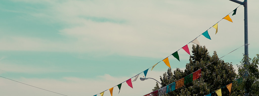 Strings of colorful festival pennant flags strung diagonally across a blue sky above green trees.