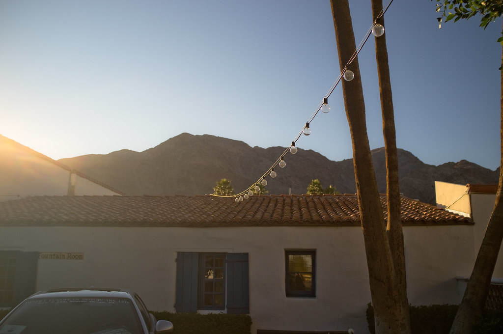 Patio lights strung beside palm trees and a Spanish villa with desert mountains during golden hour.