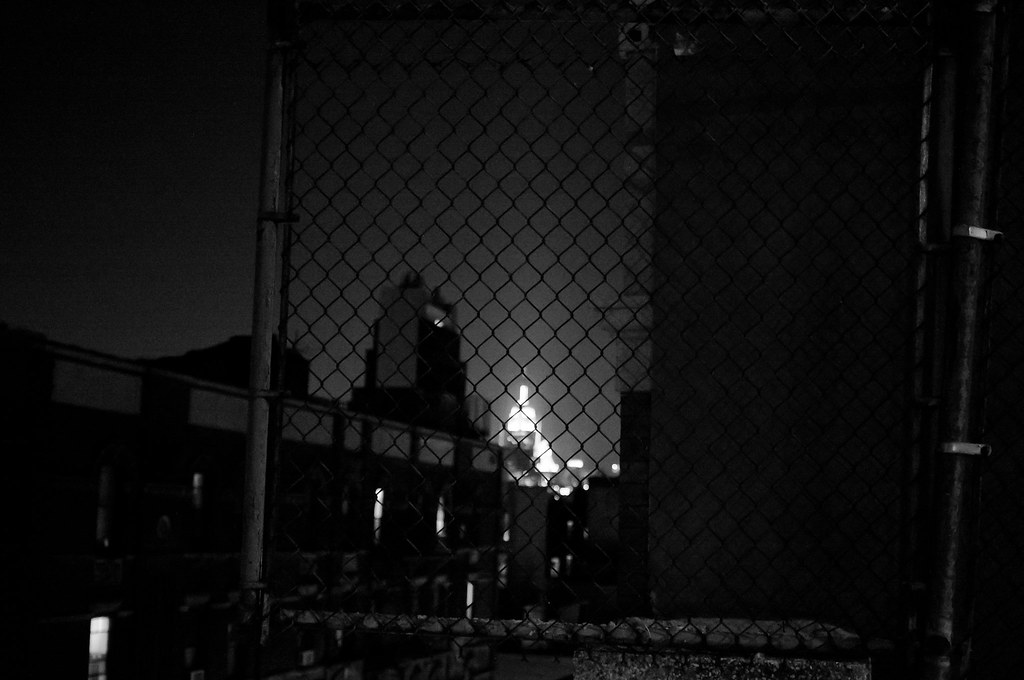 Nighttime view looking through a rooftop chain-link fence toward distant illuminated skyscraper spires.