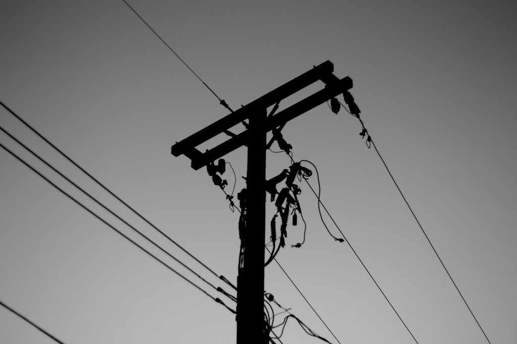 Silhouette of an electric utility pole with crossbars, insulators, and dangling wires against sky.
