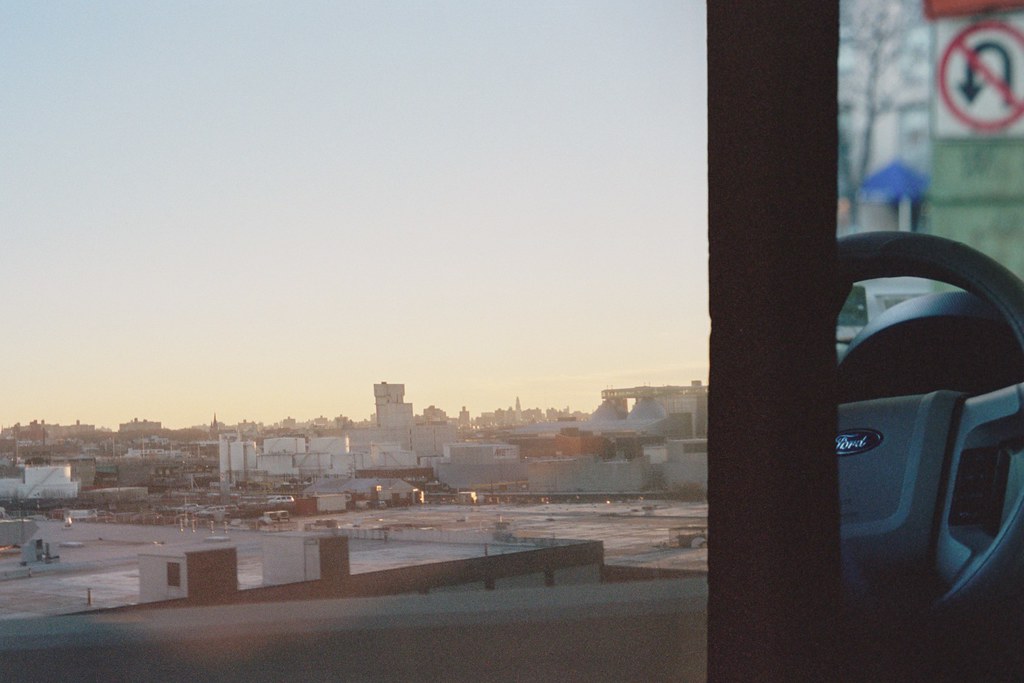 Driver's view past the steering wheel toward an industrial cityscape and skyline at dusk.