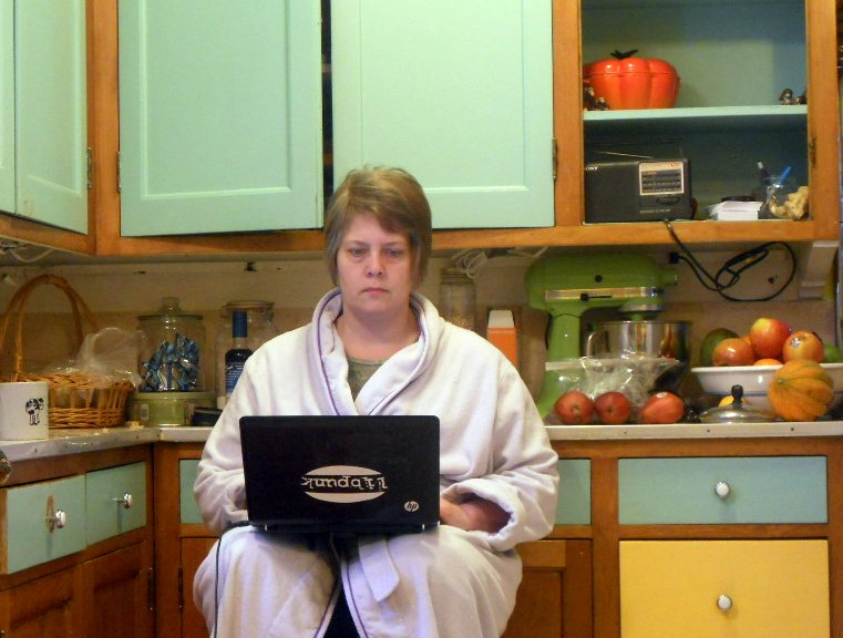 femme en robe de chambre dans sa cuisine assise sur une chaise avec un ordinateur portable sur les genoux