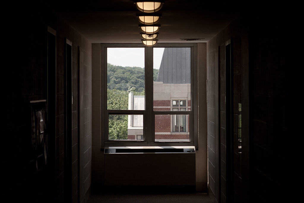 Dim hallway corridor looking toward a large window framing a sunlit brick collegiate building and trees.
