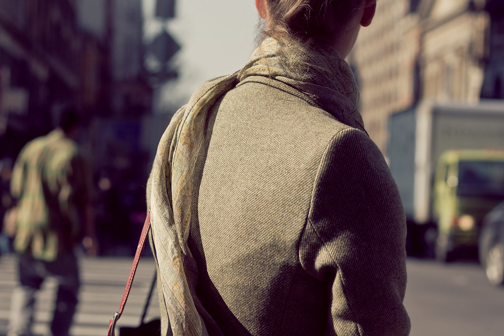 Woman with pinned hair in a tweed blazer and patterned scarf waiting at an urban crosswalk.
