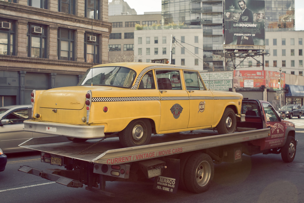 Vintage yellow Checker taxicab loaded onto a flatbed tow truck before an 'Unknown' movie billboard.