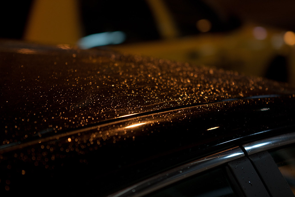 Golden streetlight reflections sparkling in raindrop beads along the curved roof of a car at night.