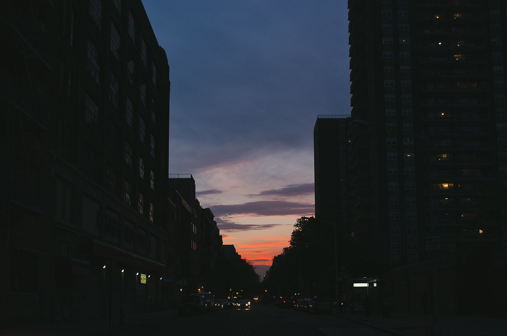 Dramatic orange sunset glow between silhouetted New York City buildings along a street at dusk.