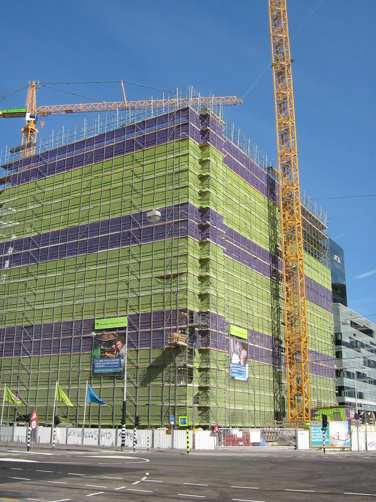 A building under construction wrapped in scaffolding and green netting, with two tower cranes standing over it and hoarding along the sidewalk.