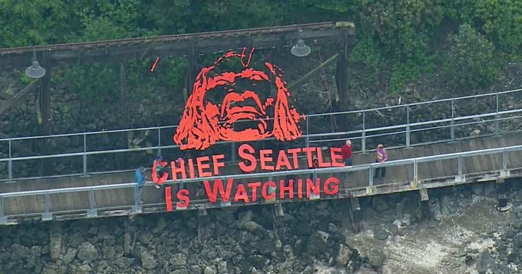 Several people putting up a net banner with an orange outline of Chief Seattle's face and text underneath the face - "Chief Seattle is Watching"