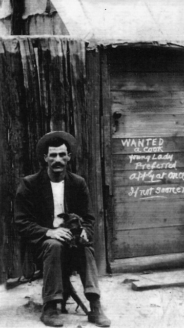 William Kelly sitting outside a hut. Sign: 