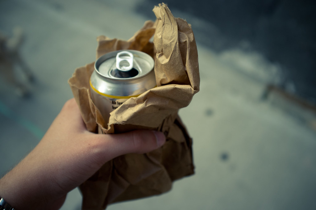 Hand holding a canned beer in a brown paper bag while walking pug dog Chino along a sidewalk.