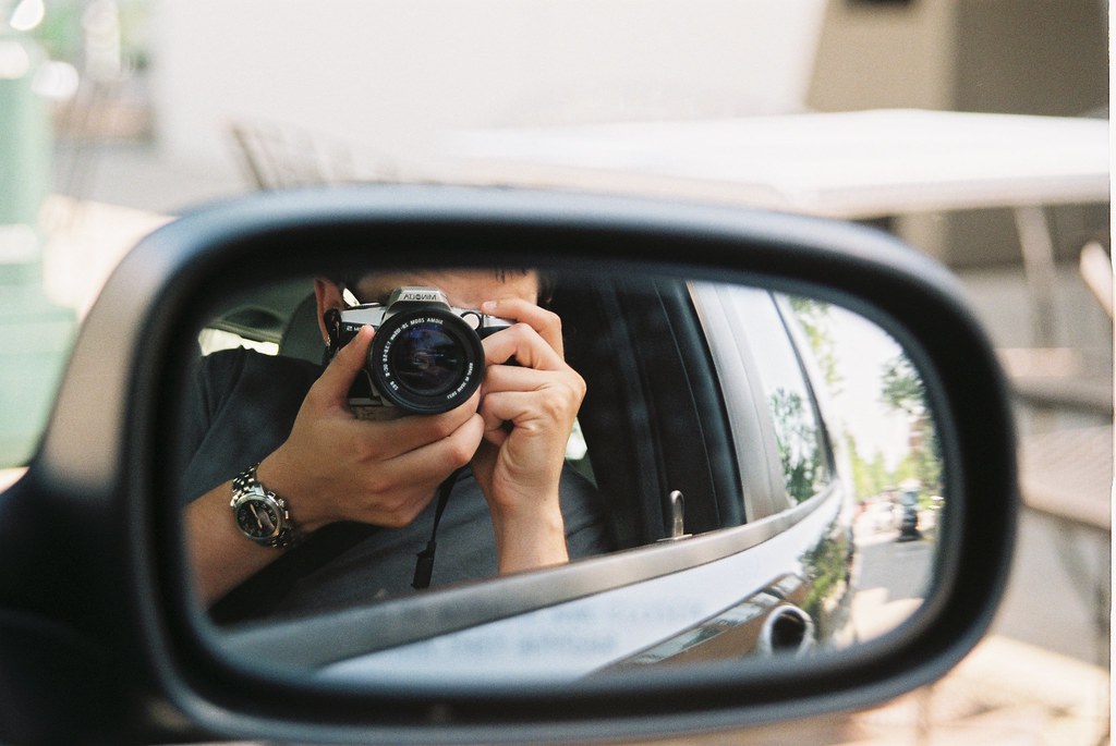 Car side-mirror reflection showing the photographer inside the vehicle holding up an SLR camera.