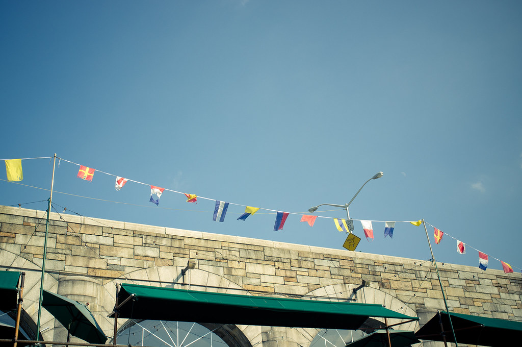 Line of colorful nautical pennants fluttering against a blue sky above stone arches and green awnings.