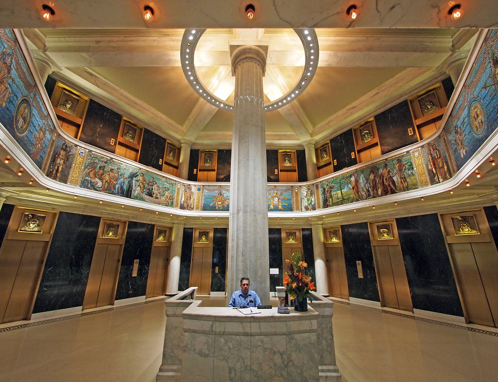 Lobby of the Marquette Building, Chicago. A column stands in the center of the dome room with two floors. There are murals on the balcony of the second floor and a man stands at the center of the photo  in the lobby. 