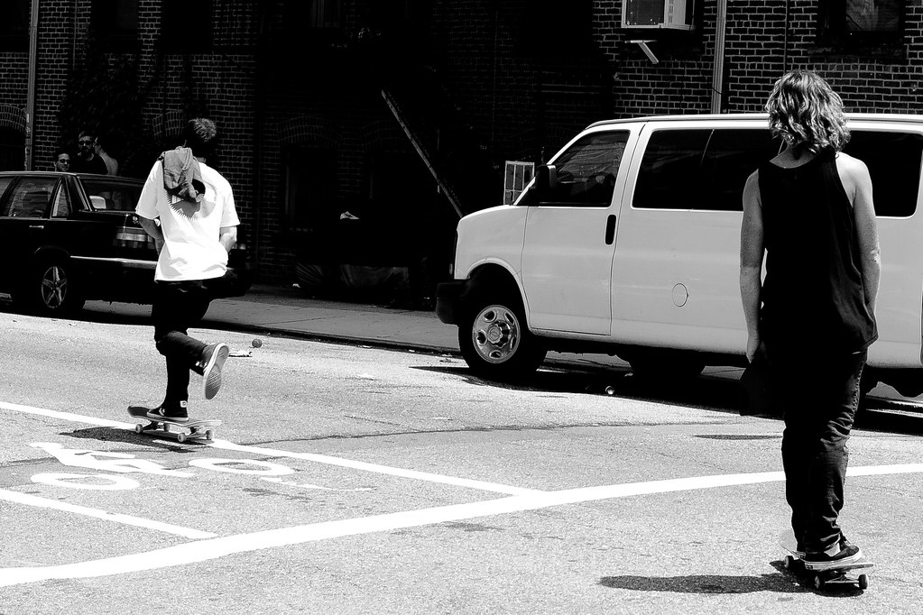 Two skateboarders pushing down a New York City street across a green bike lane past brick buildings.
