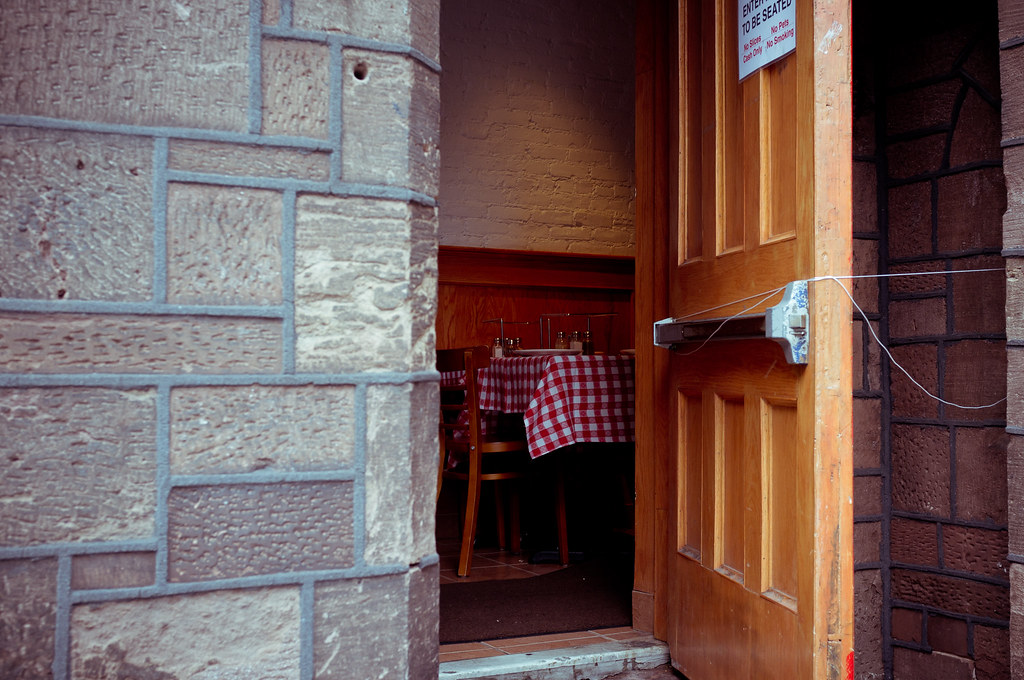 Open wooden door of a stone building revealing a dining area with red-and-white checkered tablecloth.