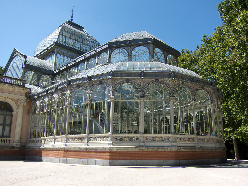 Crystal Palace in Madrid; a glass building with window arches that make up the bottom part of the building. There is a tower-like structure that sits at the top of the building (left), and a tree that sits on the right side of the photo. Moreover, blue skies occupy the background. 
