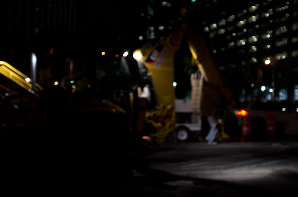 Yellow Caterpillar excavator on an urban street construction site with shimmering lights at night.
