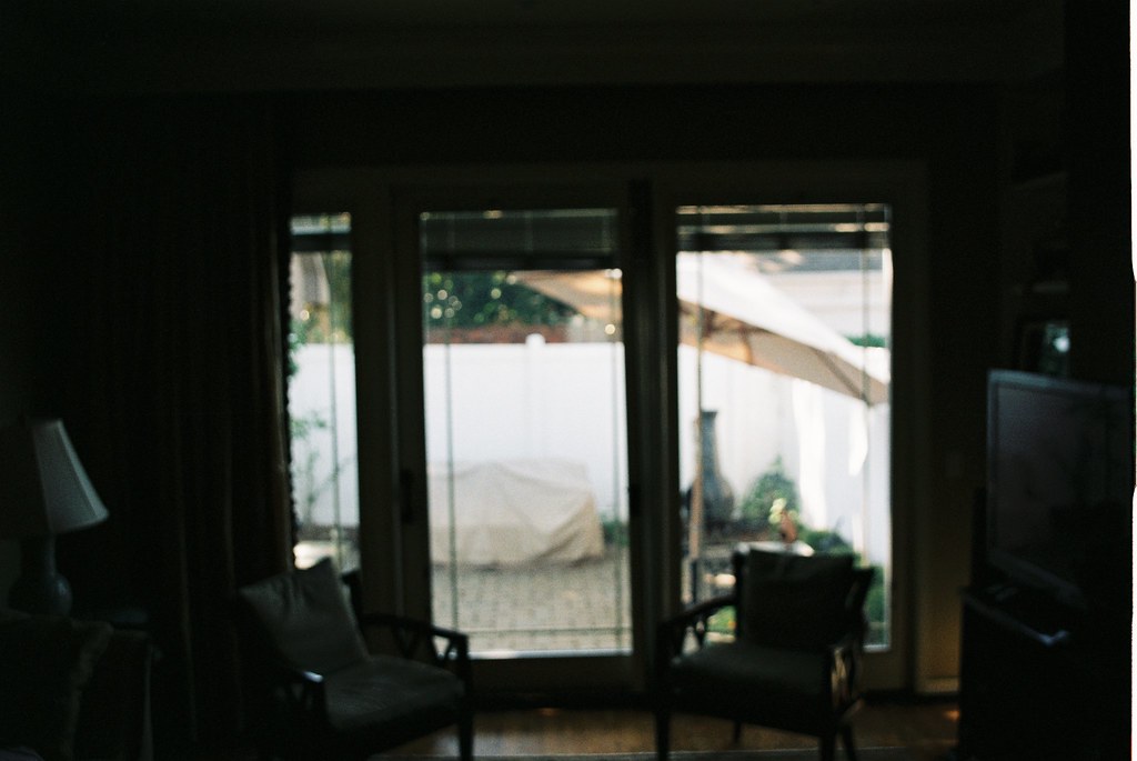 View through sliding glass patio doors toward a sunlit backyard patio with chairs and an umbrella.