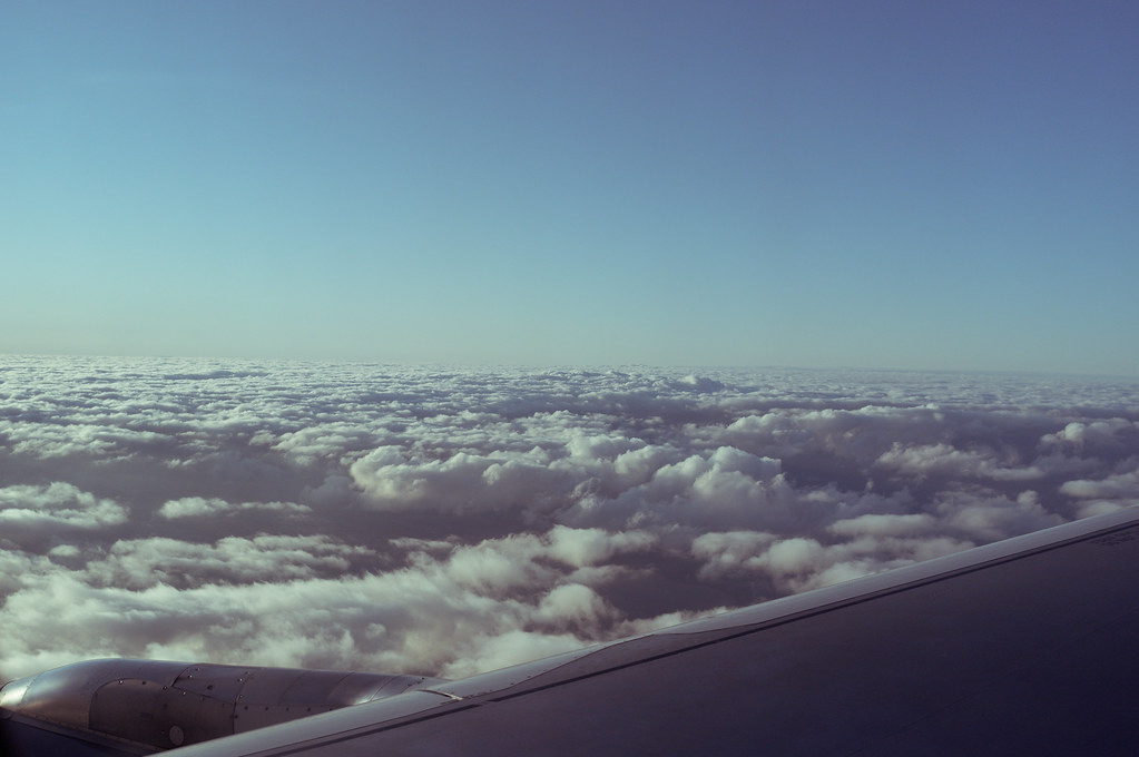 Aerial view from an airplane window over an aircraft wing above rolling white clouds and blue sky.