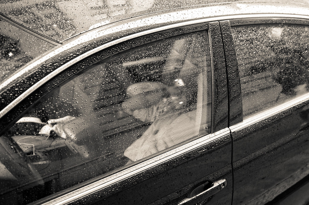 Monochrome view of rain-beaded car windows with the silhouette of a driver wearing a cap at the wheel.