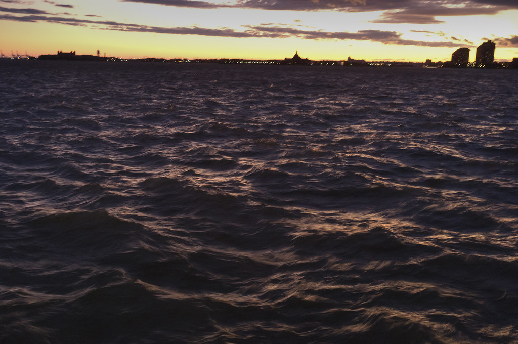Choppy dark water of New York Harbor rippling under a sunset sky with distant shoreline silhouettes.