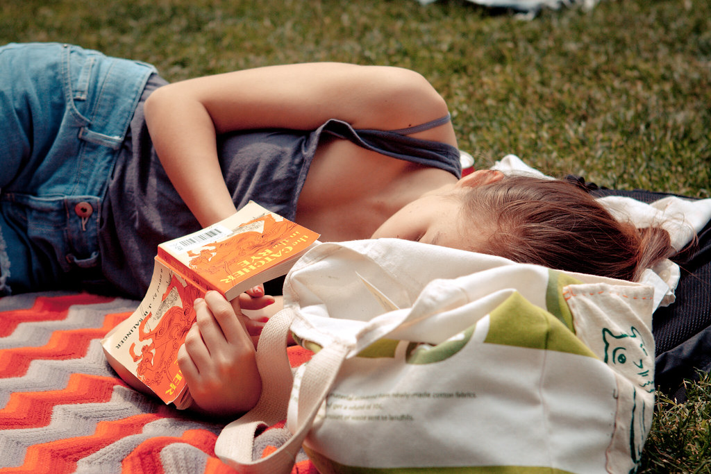 Young woman on a chevron blanket reading J.D. Salinger's 'The Catcher in the Rye' beside an open tote bag.