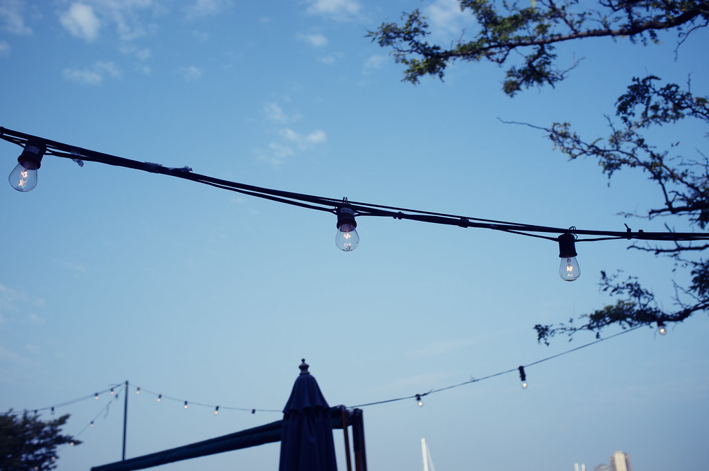 Strands of warm incandescent bistro light bulbs stretched across a clear blue morning sky above umbrellas.