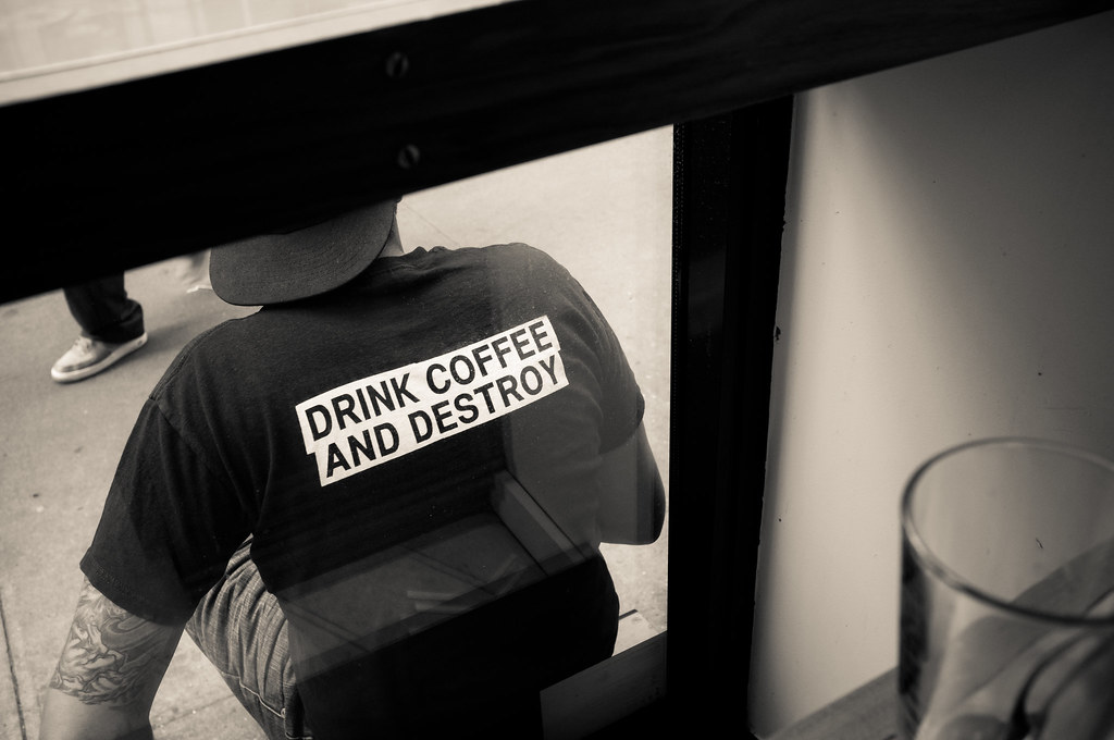 Customer sitting on a stoop in a t-shirt reading 'DRINK COFFEE AND DESTROY', seen through a cafe window.
