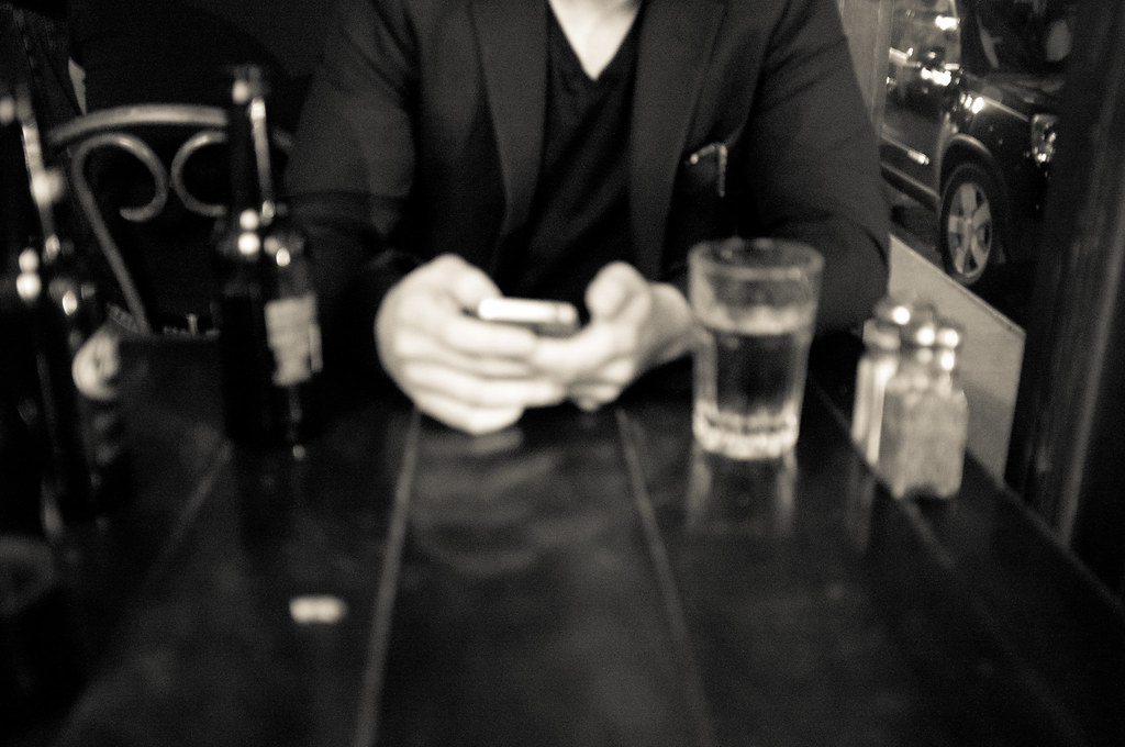 Black and white view across a bar table of Chris holding a smartphone, with beer bottles in soft focus.