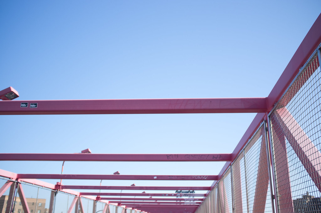 Salmon-pink steel crossbeams and wire mesh cage of a pedestrian overpass bridge framing an open blue sky.