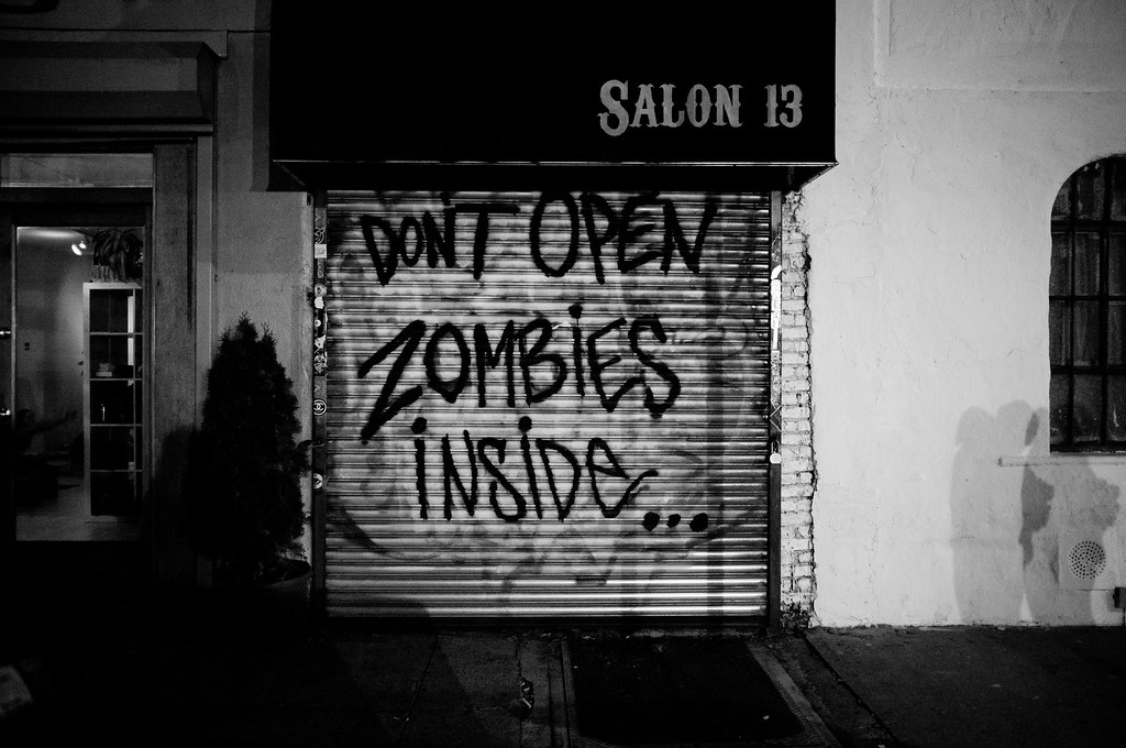 Storefront gate with graffiti 'DONT OPEN ZOMBIES INSIDE...' beneath a Salon 13 awning in New York City.
