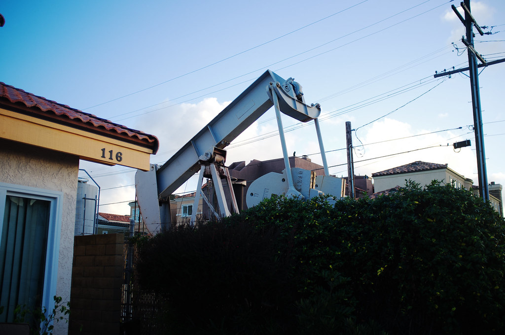 Mechanical oil pumpjack working behind dense green hedges and residential stucco homes in California.