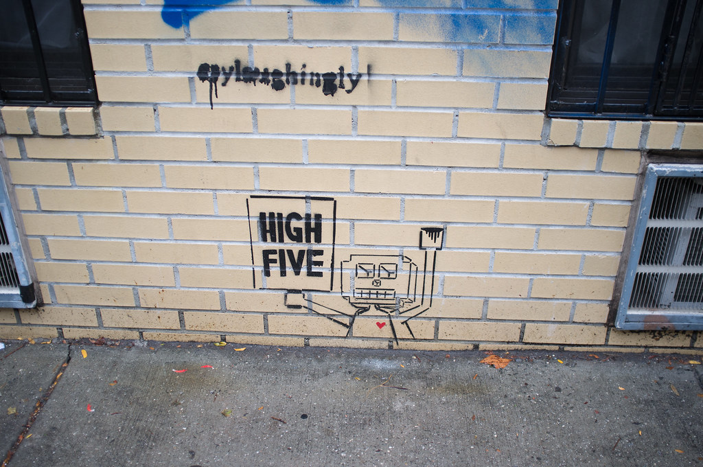 Stencil art on a yellow brick wall of a robot with a red heart holding a 'HIGH FIVE' sign.
