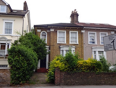 A semi-detached pair of houses with the edge of another to the left.  The house in the centre has exposed brickwork and sash windows, while its neighbour to the right has pebbledashing and modern PVC-framed windows.  Copious shrubbery grows in its front garden.