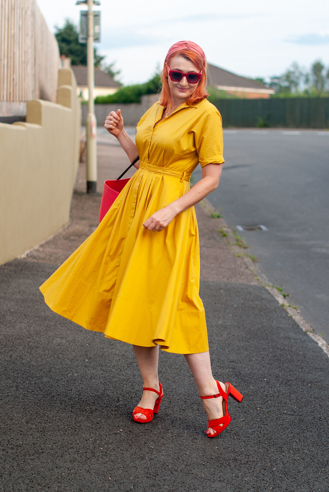 Over 40 fashion blogger Not Dressed As Lamb wearing a yellow summer dress in a shirt style \ red strappy block heels \ pink bucket bag \ pink headband \ red square sunglasses