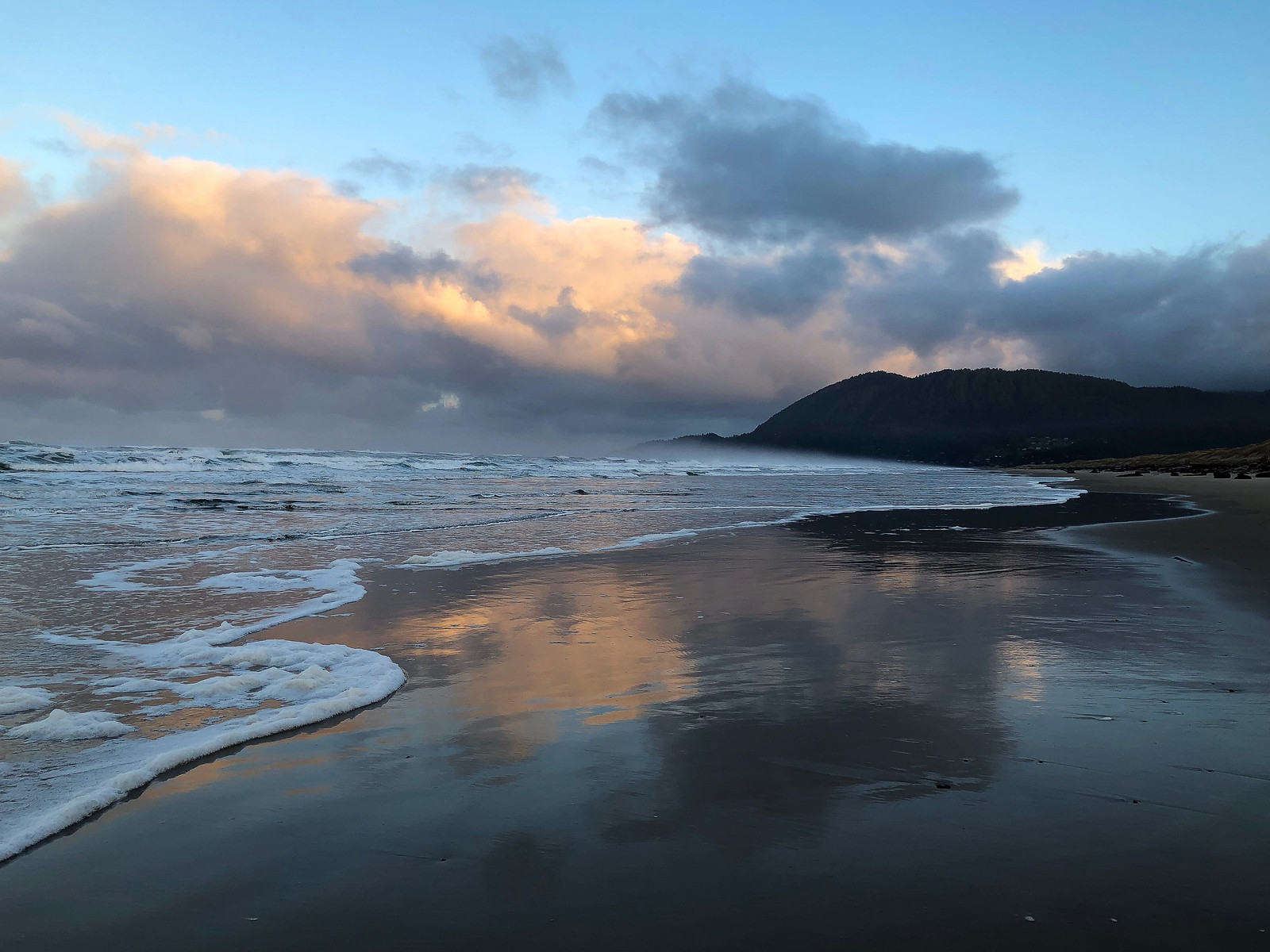 View to the north. Neahkahnie Mountain on the right background, surf and swash stretching to the left. Gray and peachy morning clouds reflected in the mirror.