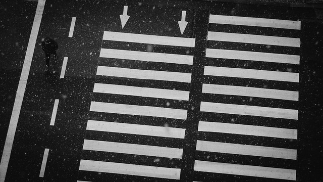 Snow falls over an urban zebra crossing viewed from above; bold white stripes contrast with dark asphalt, with a lone pedestrian near the left edge.