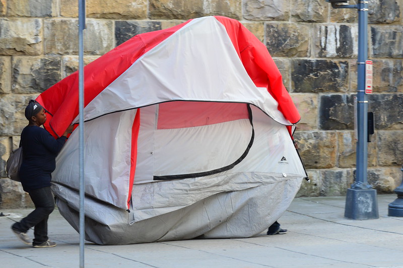  A woman holds and carries one side of a red and gray tent. The bottom sags and drags on the ground, indicating there are belongings inside.