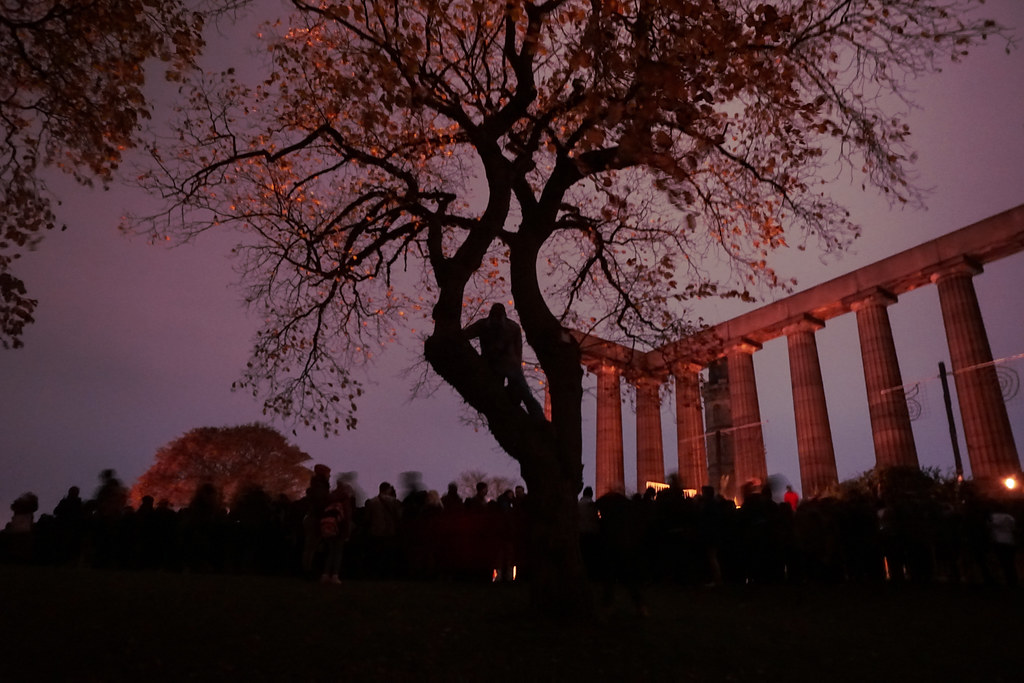 A silhouetted figure in a tree above a crowd and Acropolis at Samhuinn Fire Festival