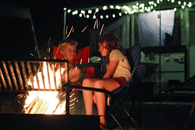 a boy and girl sit facing each other talking at a campfire with rv in background