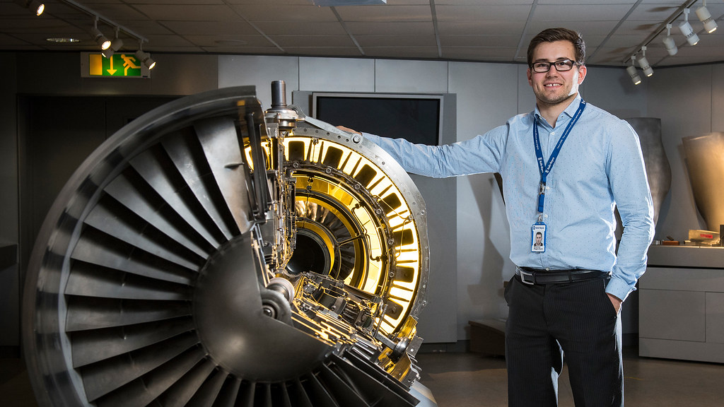 A male student on placement stands confidently next to a rolls-royce engine