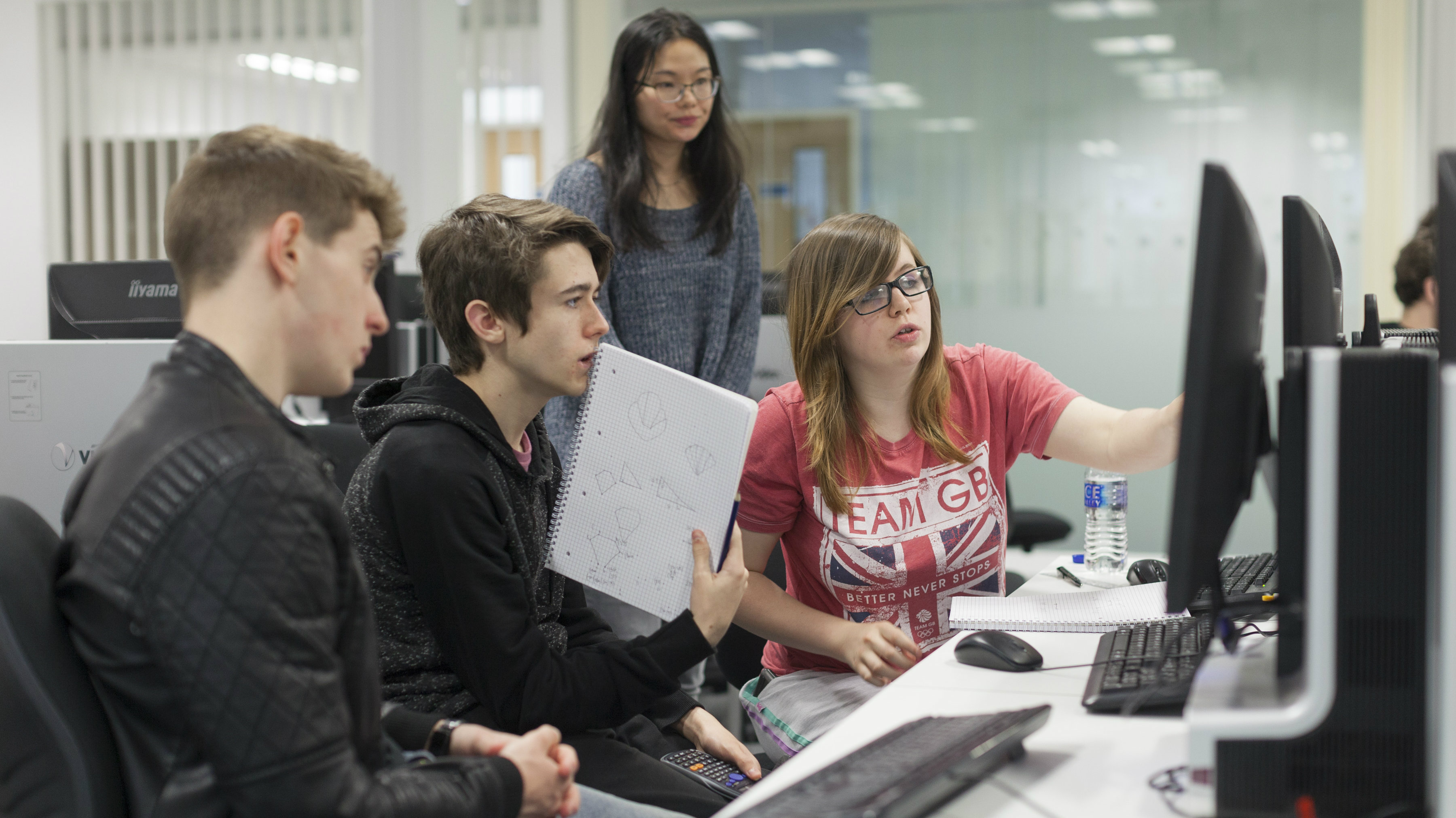 Two female and two male students huddled around two computer screens. The two young men, both of whom are wearing black, are sat down next to a young lady wearing a red Team GB t-shirt. The other young lady is stood up wearing a blue long-sleeved jumper.
