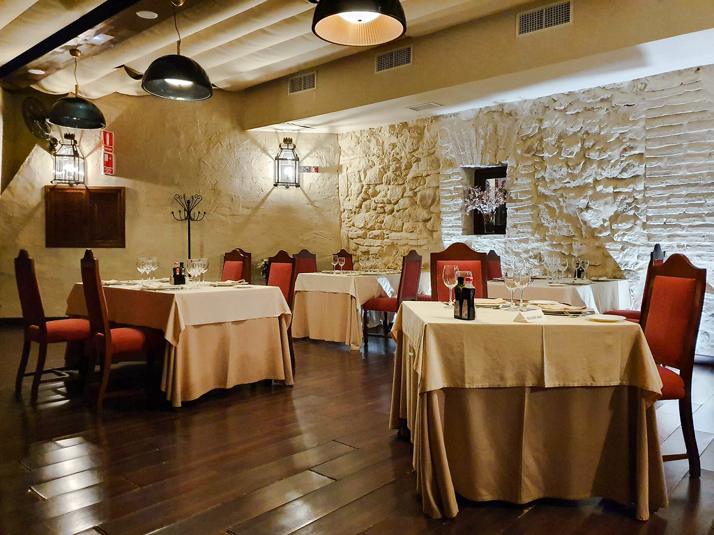 The dining hall at the hotel's restaurant. Rectangular tables covered with white table cloths, with red chair around them. The wall behind is made out of exposed bricks and stone  