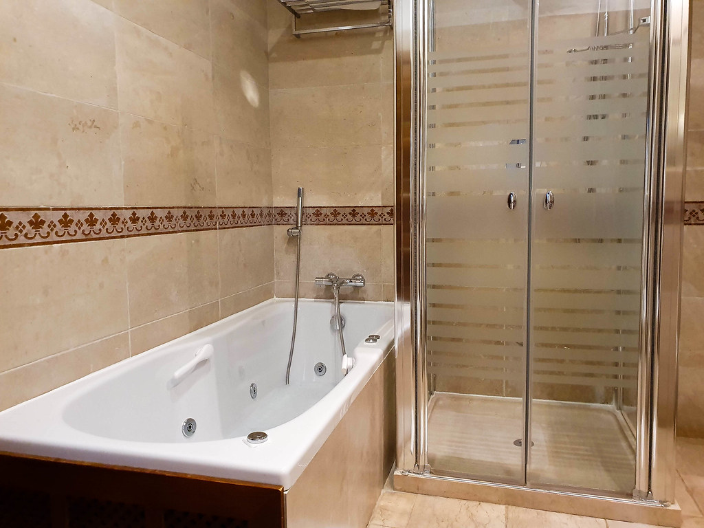 A modern bathroom, with a tub with jacuzzi on the left, and a shower on the right. The tiles on the walls are light creamy color, with a band of ornamental tiles going through the middle. 